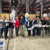 People lined up behind ribbon inside warehouse with man holding large scissors after cutting ribbon
