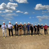 Several people stand in a row wearing hard hats and holding shovels