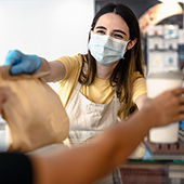 woman in medical mask behind restaurant counter handing customer a to-go bag