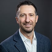 Headshot of man smiling in professional collared shirt and jacket against dark grey background