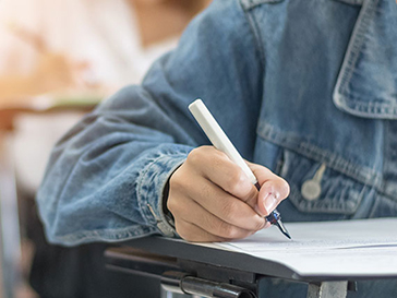 Close up of the right side of a person sitting at a table, writing on a piece of paper while wearing a jean jacket
